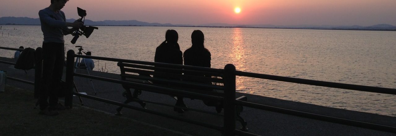写真：海に沈む夕日をベンチに座って見ている女子高生2人を後ろからカメラマンが撮影している様子